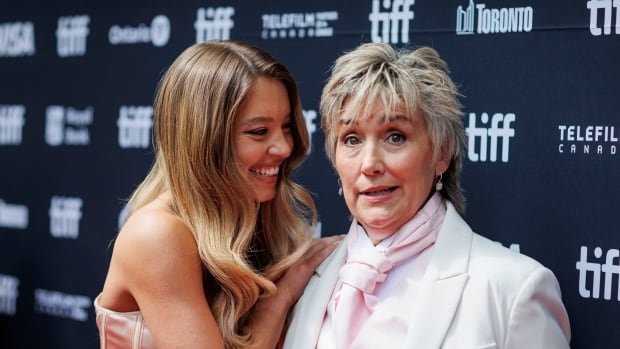 Two women holding onto one another, posing on a red carpet. One is wearing a strapless evening gown and the other a white tux with pink vest and tie.