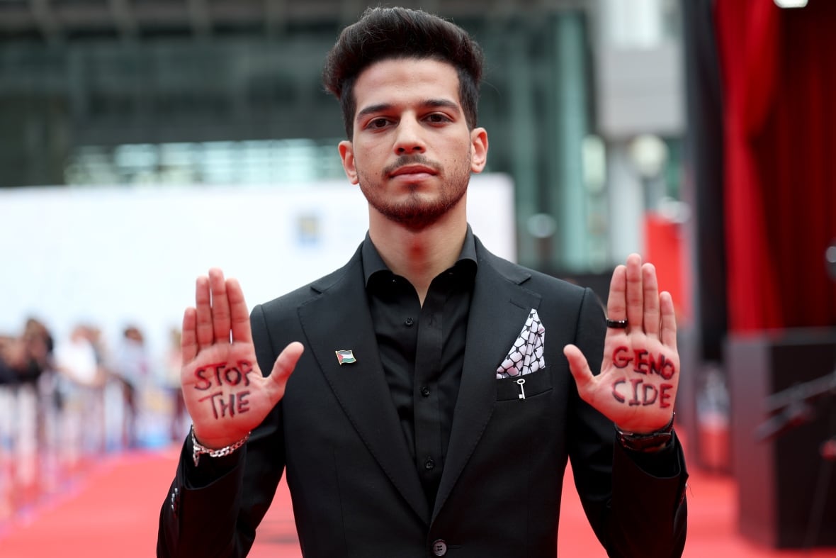 A man in a black suit stands on a red carpet at a film premiere with his hands held up facing the camera and the words "Stop the genocide" written on his palms.