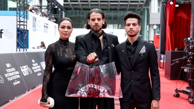 Three people standing on a red carpet at a film premiere. From left to right: A woman in a black gown, a man in a black suit holding up a plastic bag dripping with fake blood and containing a camera and a black and white print scarf; another man in a dark suit. 