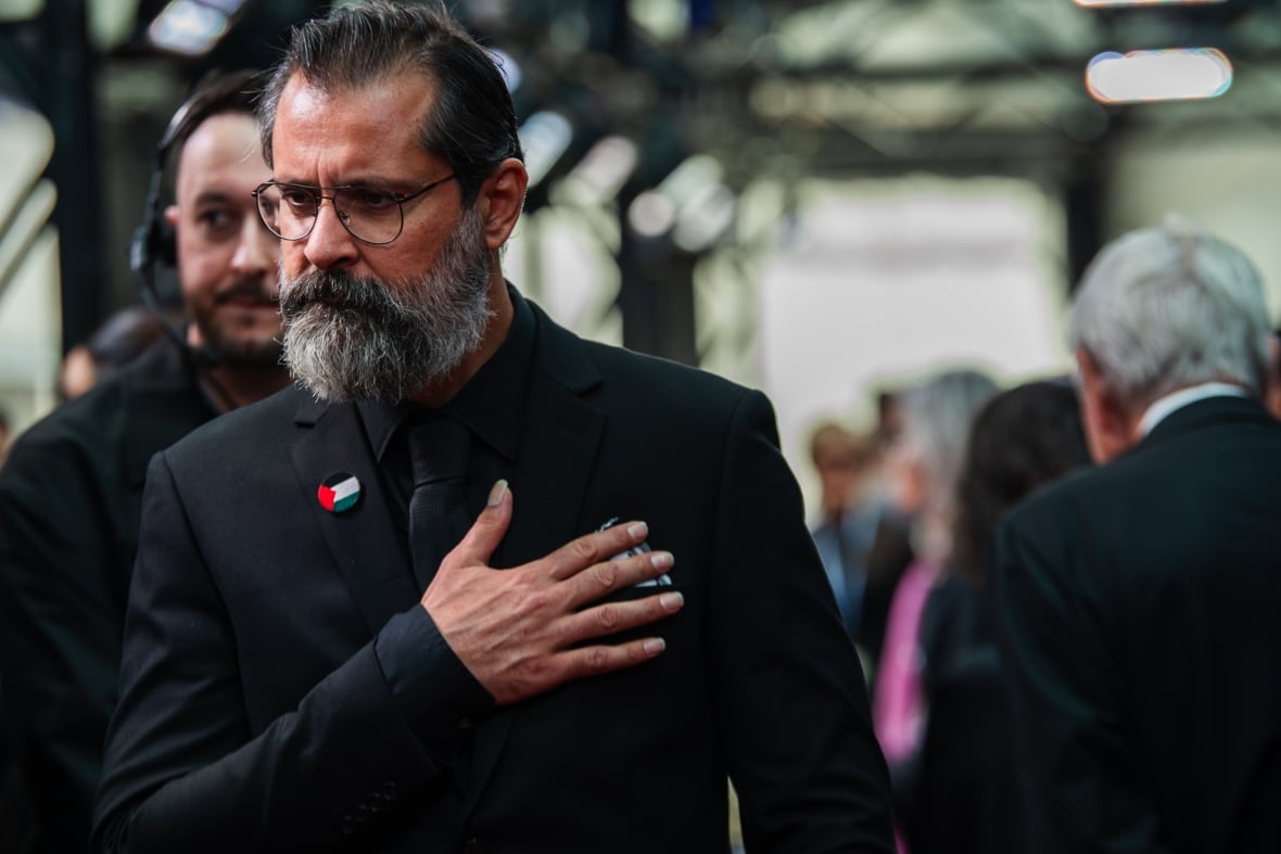 A bearded man wearing glasses and a black suit with a black, white, green and red Palestinian flag pin on his lapel. He's holding his right hand over his heart.