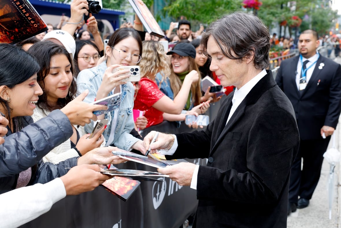 A man signs autographs to a group of people standing behind a barricade.