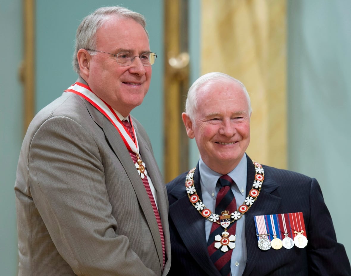A tall man with gray hair with glasses and a gray suit, with a medal hung around his neck, shakes hand with a white hair man with a dark suit showing medals.