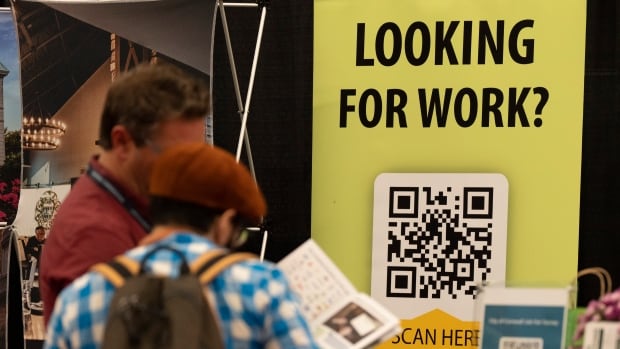 Two men hold a paper while a "looking for work" sign hangs in the back