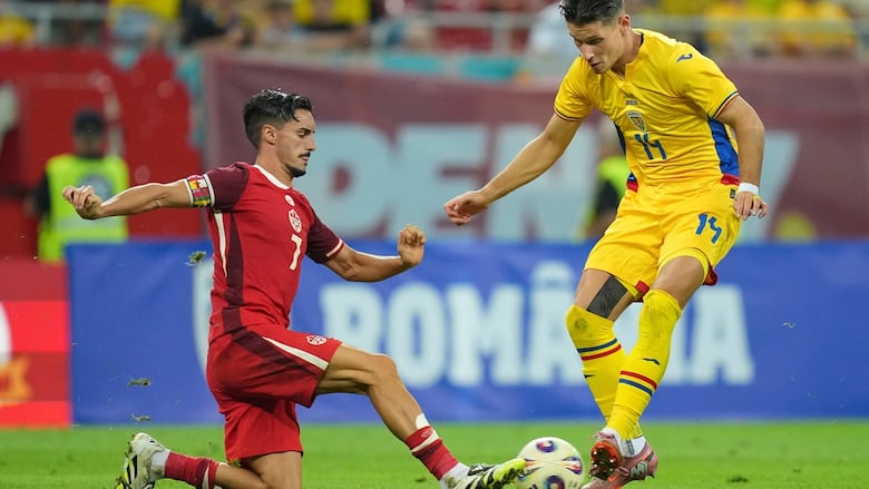 A male soccer player representing Canada lunges forward with his right leg to kick the ball away from a Romanian player.