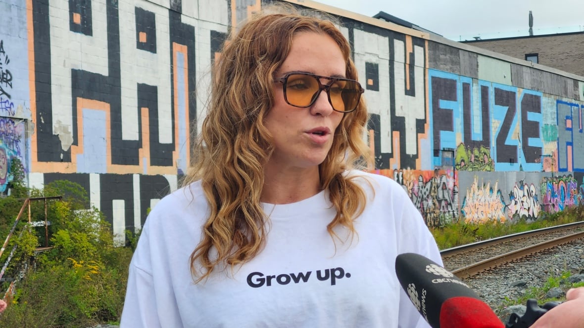 A woman with curly blonde hair stands in front of a mural that reads Pat Stay. She is wearing a white t-shirt with the words Grow Up on it.