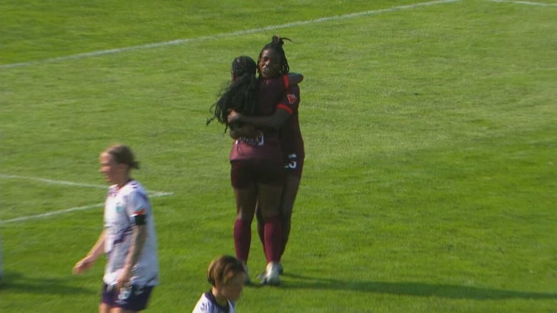 Two female soccer players hug in celebration on the field during a day game.