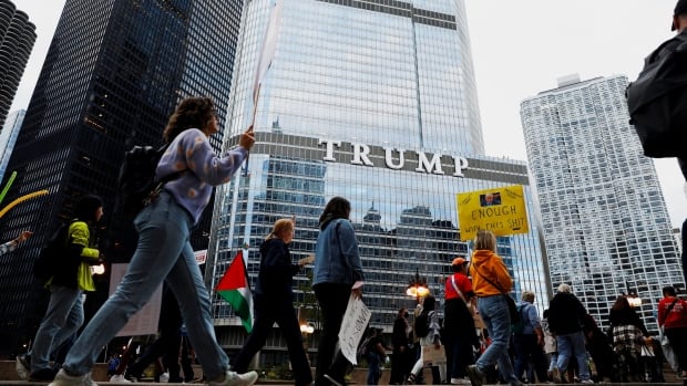 People can be seen marching and holding protest signs in front of a tall glass building with the world trump on it
