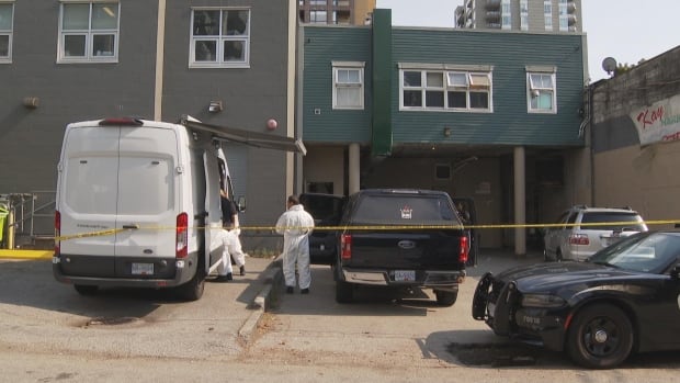 People in white 'hazmat'-type suits stand beside cars in front of a taped off apartment building beside police cars.