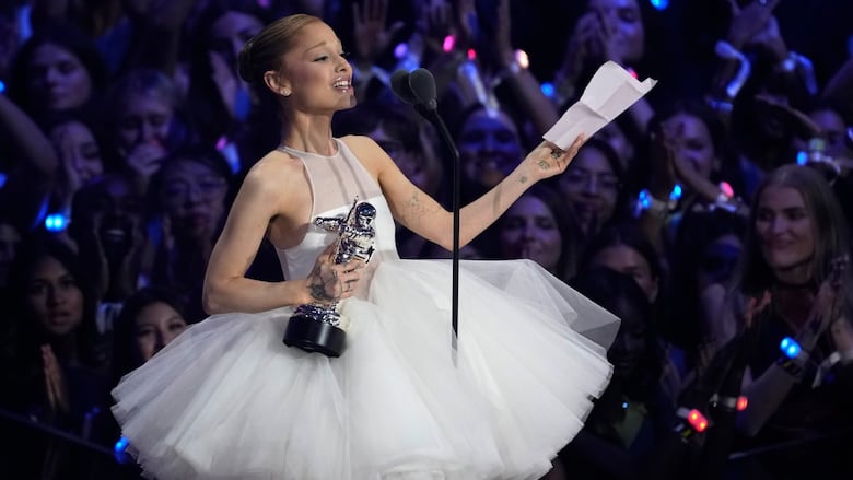 a woman in a white dress with a puffy tutu speaks into a microphone while holding a chrome spaceman trophy in one hand and a piece of paper in the other. behind her a large crowd is visible