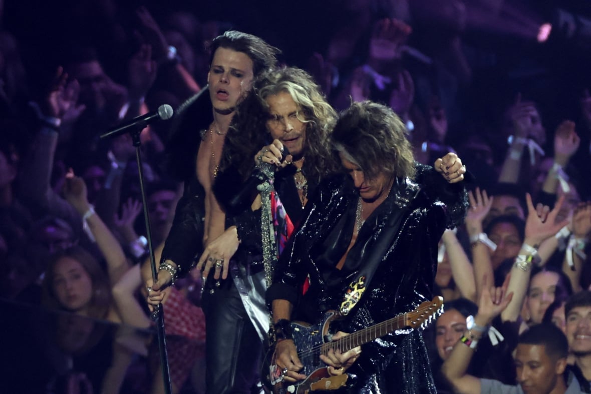 three rockstar-looking men perform on stage huddled with their arms around one abother. two on the left are singing and the third on the right is playing guitar. a crowd is visible behind them