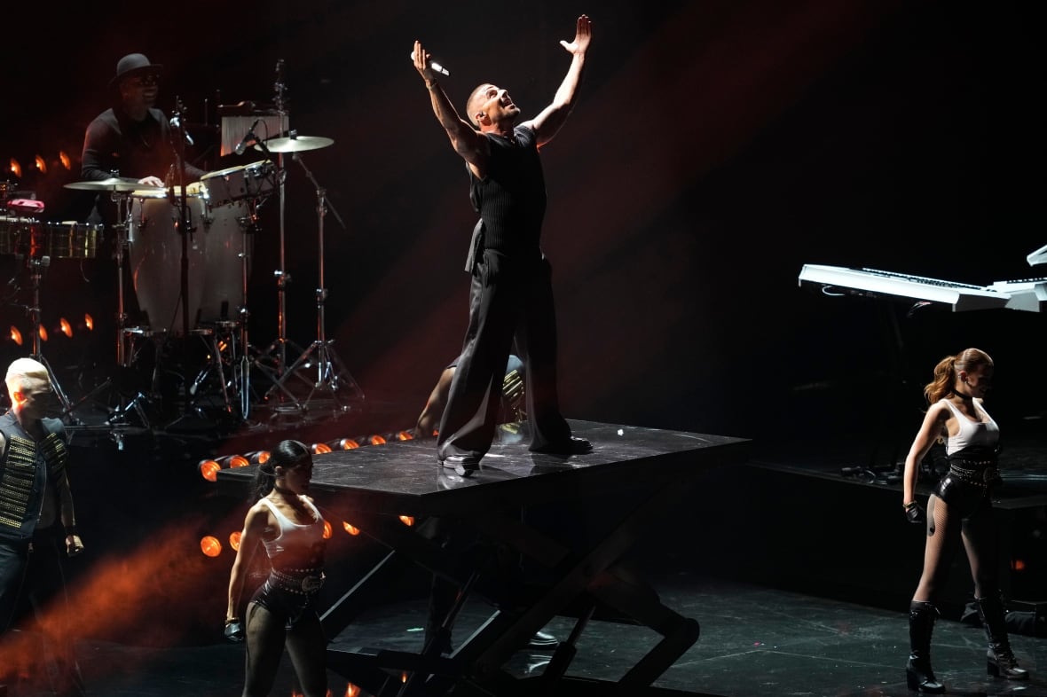 a man in black clothes holds his arms up. hes standing on a platform on a stage and holding a microphone in his hand. backup dancers can be seen around him, performing alongside him