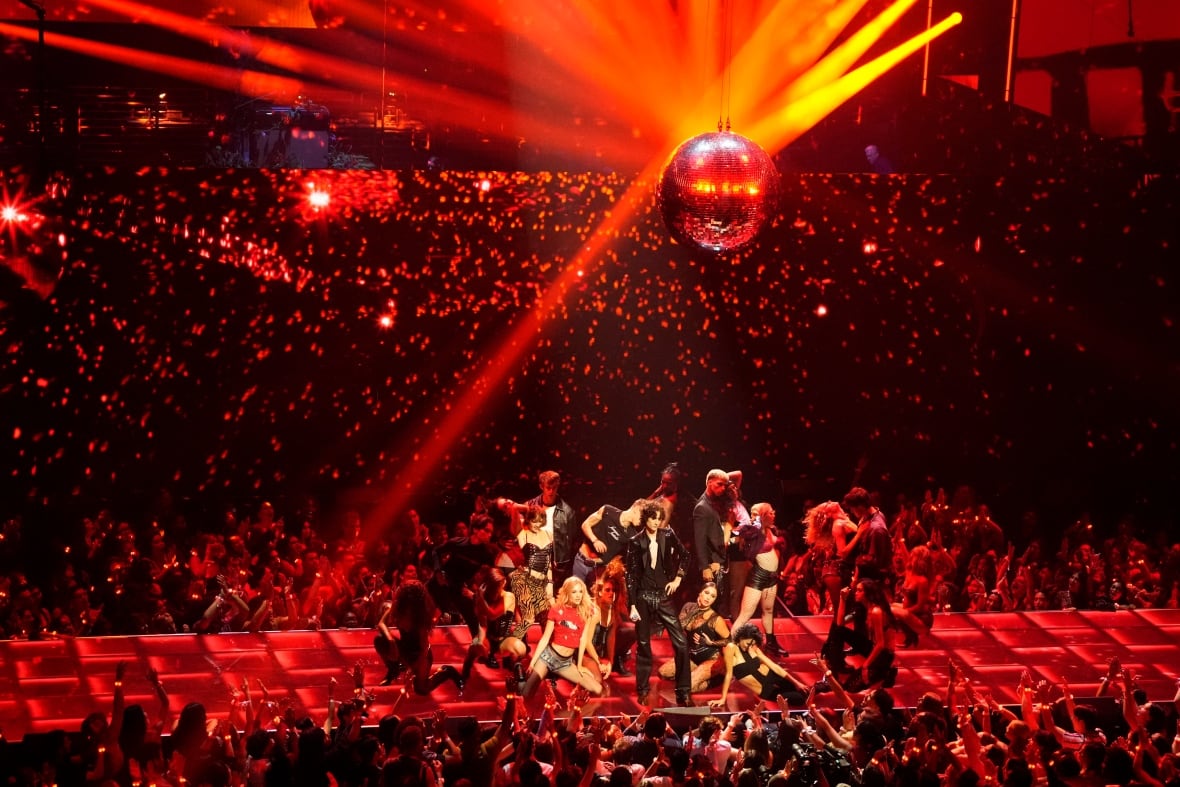 a troupe of dancers perform alongside a musical act on a stage under red lighting and a massive disco ball, as glitter rains down. a crowd surrounds the stage