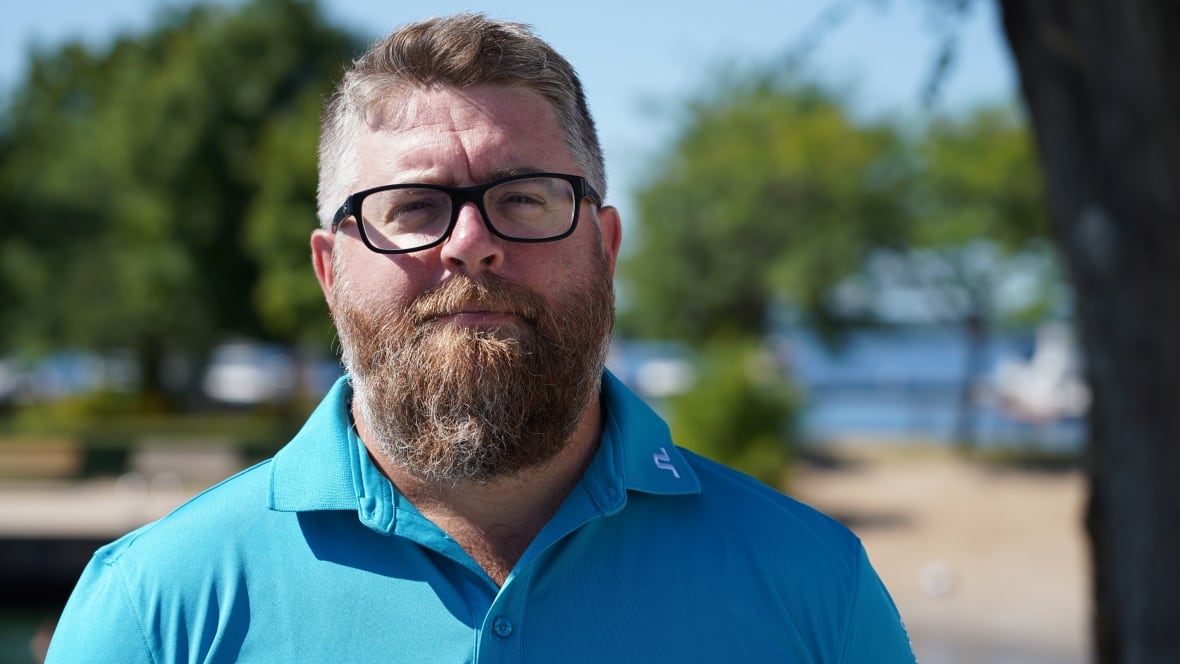 A man with reddish, gray hair and glasses stands outside at a park on a sunny day.