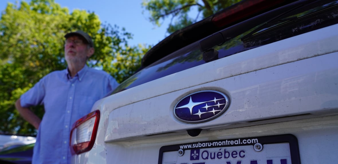 A man in a blue shirt and cap stands next to a white Subaru with a Quebec licence plate.