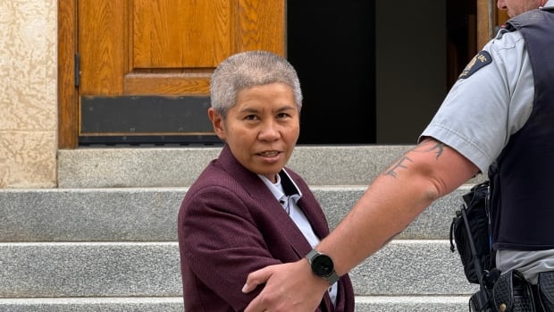 A woman is shown wearing a red suit. The arm of an officer is shown holding her arm as he pulls her up the stairs. 