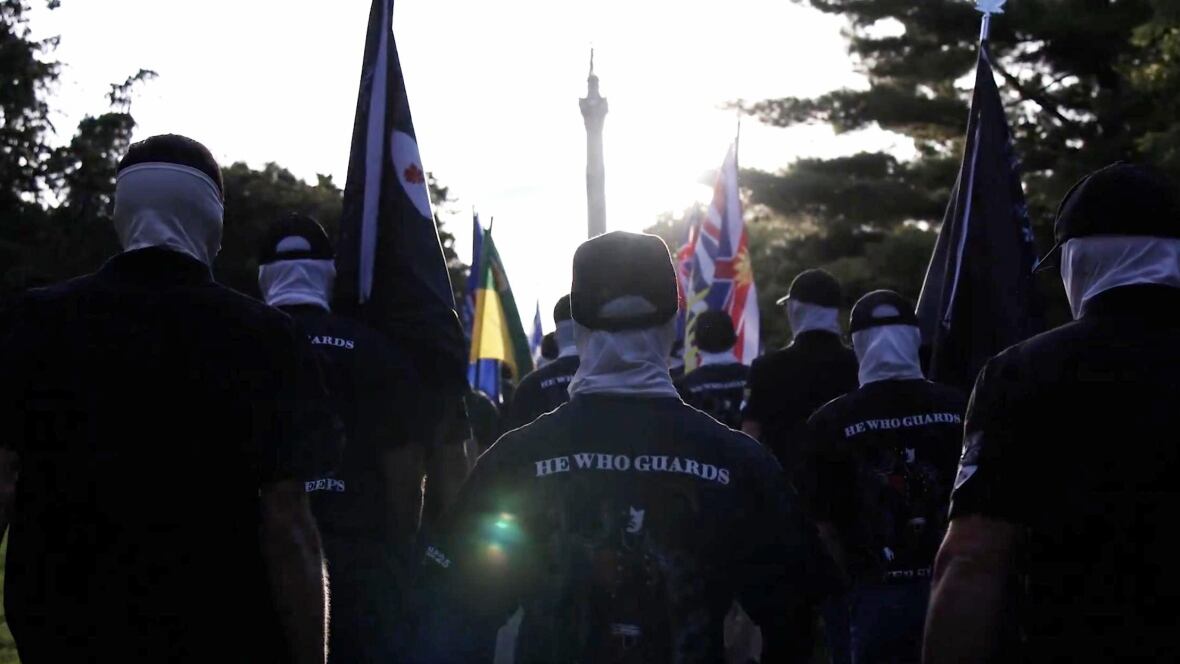A group of people masked and wearing black T-shirts with the words "He who guards" on the back, march together.