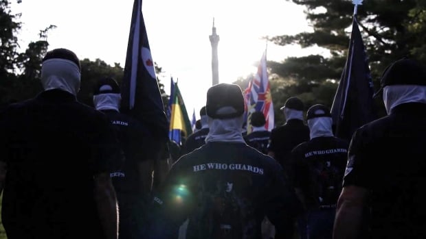 A group of people masked and wearing black T-shirts with the words "He who guards" on the back, march together.
