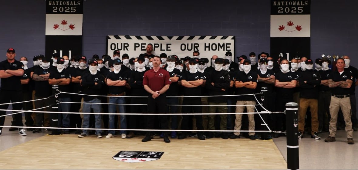 A group of mostly masked men on matching black t -shirts are published behind an indoors fighting ring.