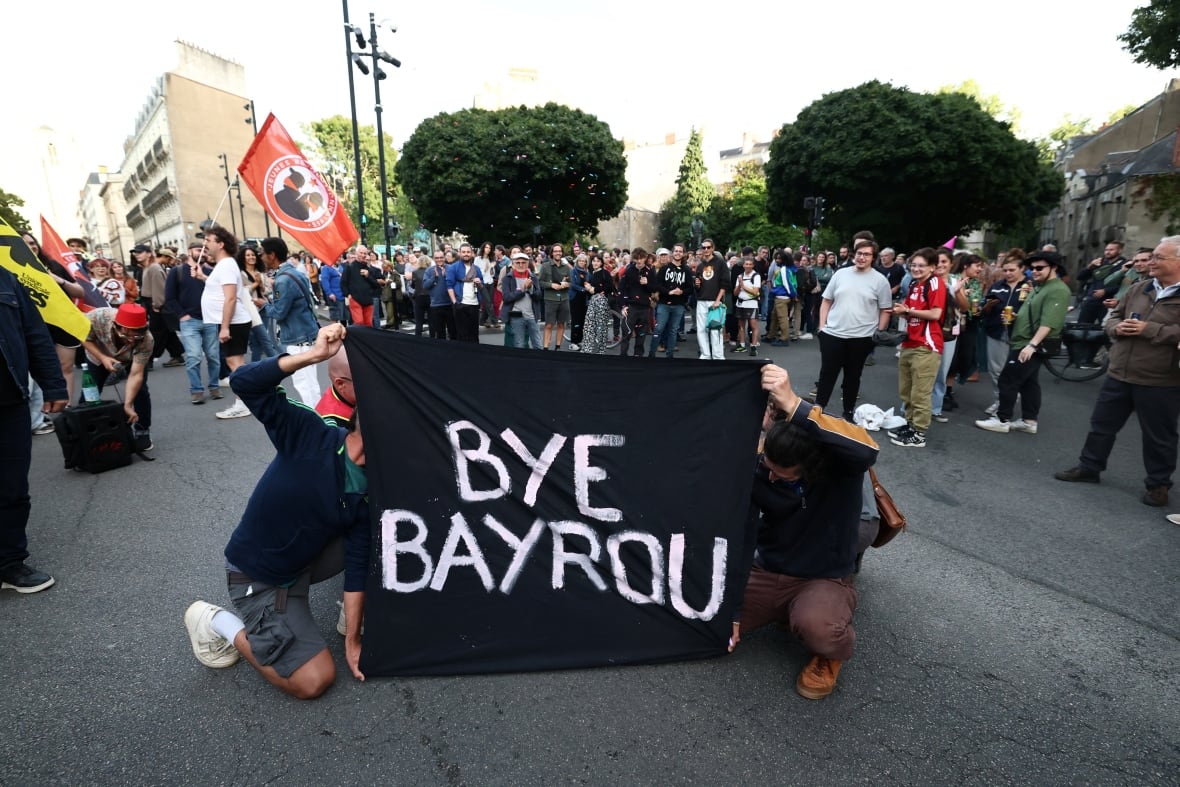 People hold a black panel with a white lettering that reads: Bye Bayrou, while people gather in the street behind them