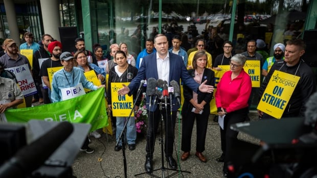 A man stands speaking at a podium, surrounded by picketers. 