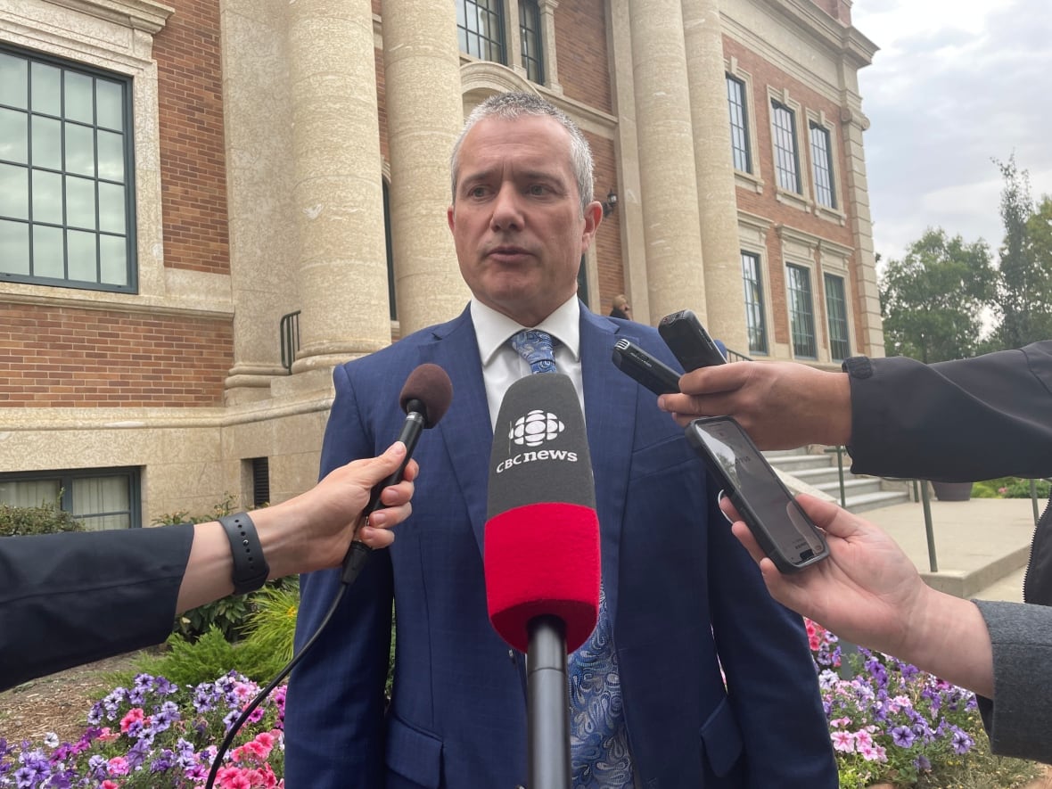 A man wearing a blue suit stands in front of a brick building as he speaks into four microphones being held by reporters.