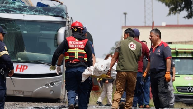 Rescue workers remove a body at the scene of a deadly accident where a freight train slammed into a double-deck bus in Atlacomulco, Mexico, Monday, Sept. 8, 2025. A group of men carry a body wrapped in white down a road strewn with debris. A bus with the top and one side smashed up is visible at the left side of the image.