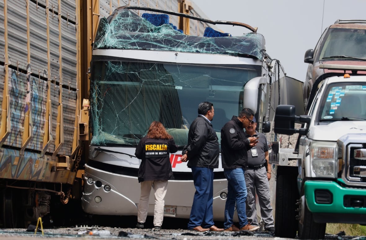 A broken bus is crushed on the side of the train, and its upper and sides crash. A few people stand in front of the bus.
