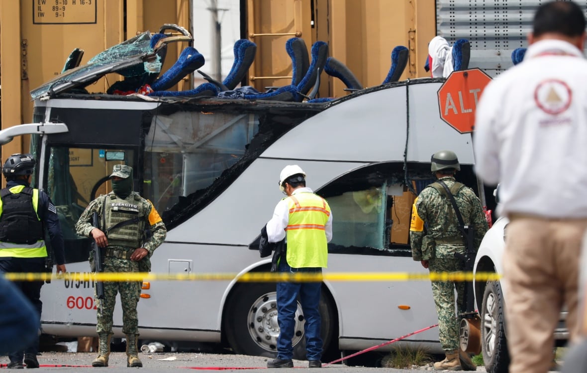 Behind a clear line of the yellow tape, the bus is visible next to the train. The bus was severely damaged from the top, with flat seats near the front. A few people stand, including a couple wearing military uniform.