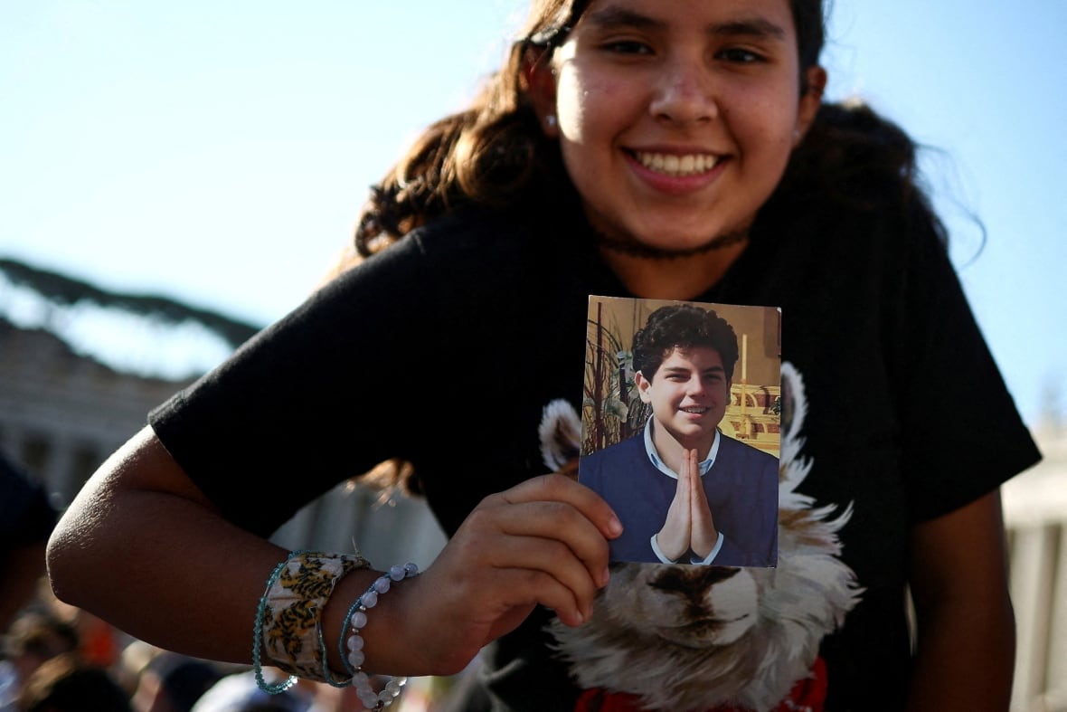 A smiling teenage girl bearing a picture of a teenage boy with curly hair smiling and making the hands of prayer