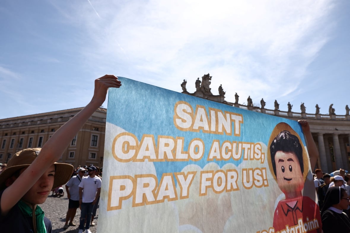 People carry a banner with a Lego man with curly brown hair and words "The sharp St. Carlo pray for us!" In the big bubble messages. 