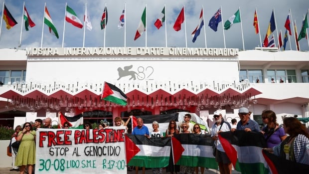 people stand holding palestinian flags and a big sign that says "free palestine, stop al genocidio". behind them, the building that houses the Venice Film Festival can be seen