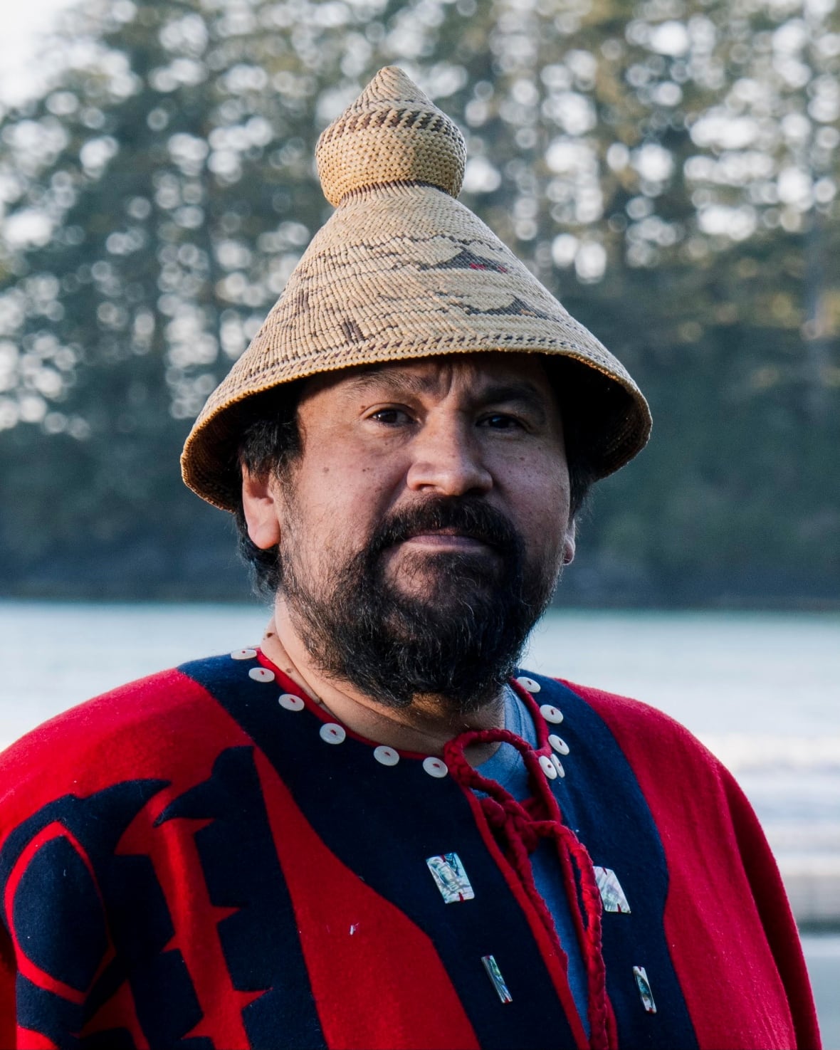 Photo is a portrait of an indigenous man wearing a woven hat and a cape with buttons and coastal art.