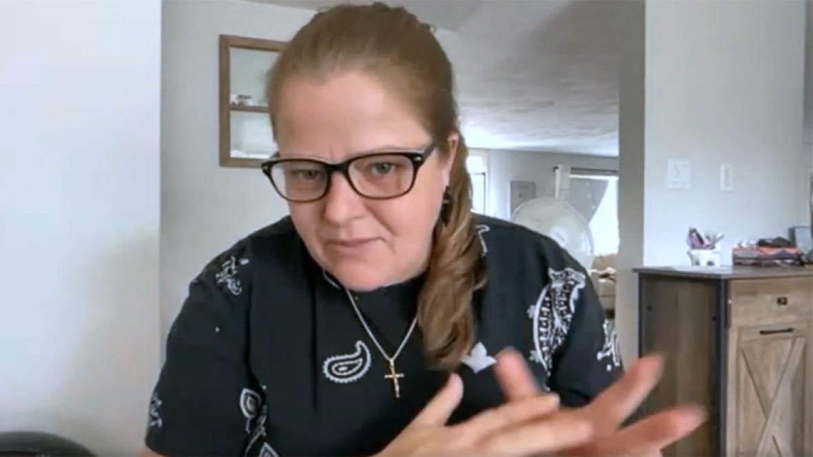 A woman with brown hair tied back, wearing a black and white top and black glasses, gesturing with her hands, while talking during a video call at her home.