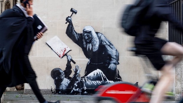 a wall mural depicts a judge beating a protester who has fallen to the ground with a gavel. on the street and sidewalk in front of the wall, a judge talks on the phone and walks, and a cyclist speeds by on a bike