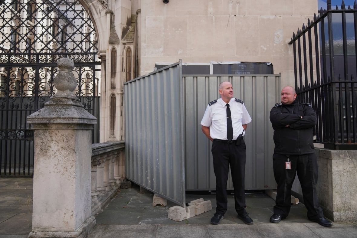 in front of an old, gothic building, two security guards stand on guard. behind them, a spot on the wall where a mural is painted is covered completely by black plastic taped to the wall, and metal barriers.