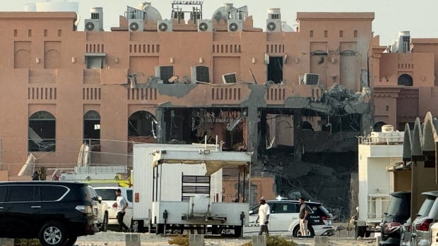 Three people walk in front of  a destroyed building with police vehicles parked in front of it.