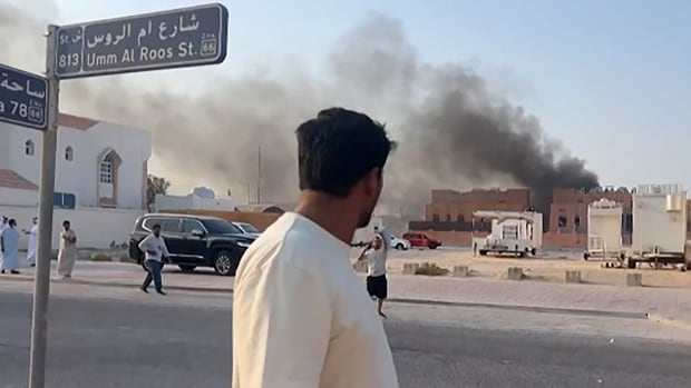 Man stands beside a street sign looking at a low-rise building with smoke billowing from it, and a handful of other people walking in the distance.