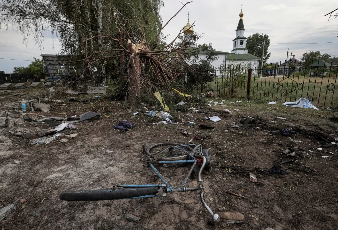 A severe damaged tree and a bike appears on the dirt ground at the front of the image, with a church -like building in the background.