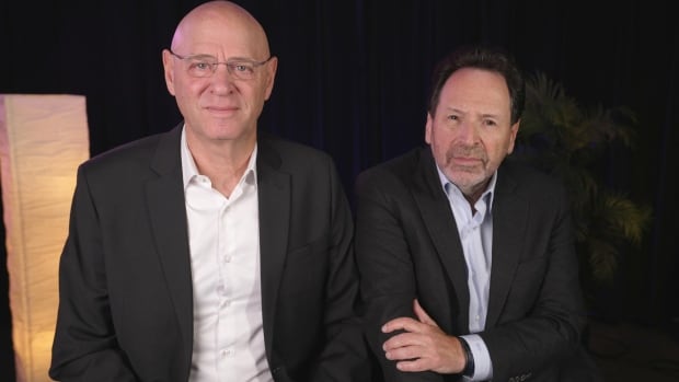 Two men sit side by side and give small smiles at the camera, both in casual suits. 
