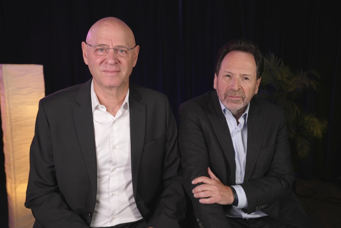Two men sit side by side and give small smiles at the camera, both in casual suits. 