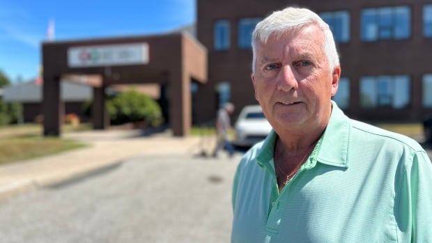 A man with white hair and a turquoise golf shirt smiles at the camera in front of a brick building on a sunny, summer day. 