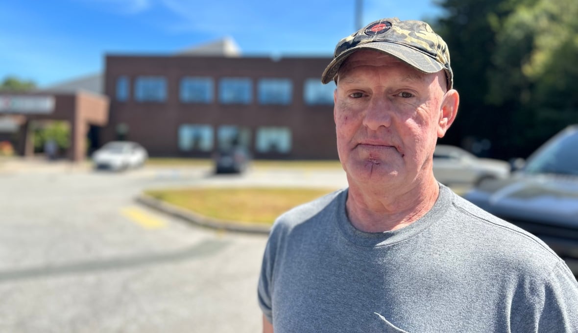 A man with a camouflage cap and a grey short-sleeve t-shirt looks at the camera with a serious face, while standing outside a brick building on a sunny, summer day. 