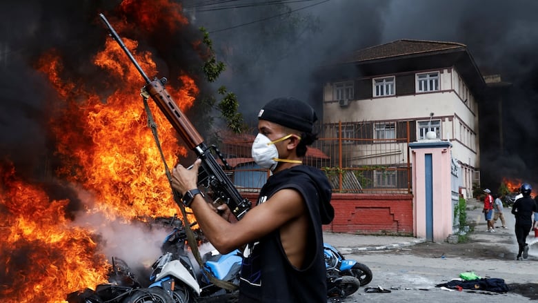 A masked man brandishes an assault rifle on a street. The tangled remains of one or more vehicles burn behind him.