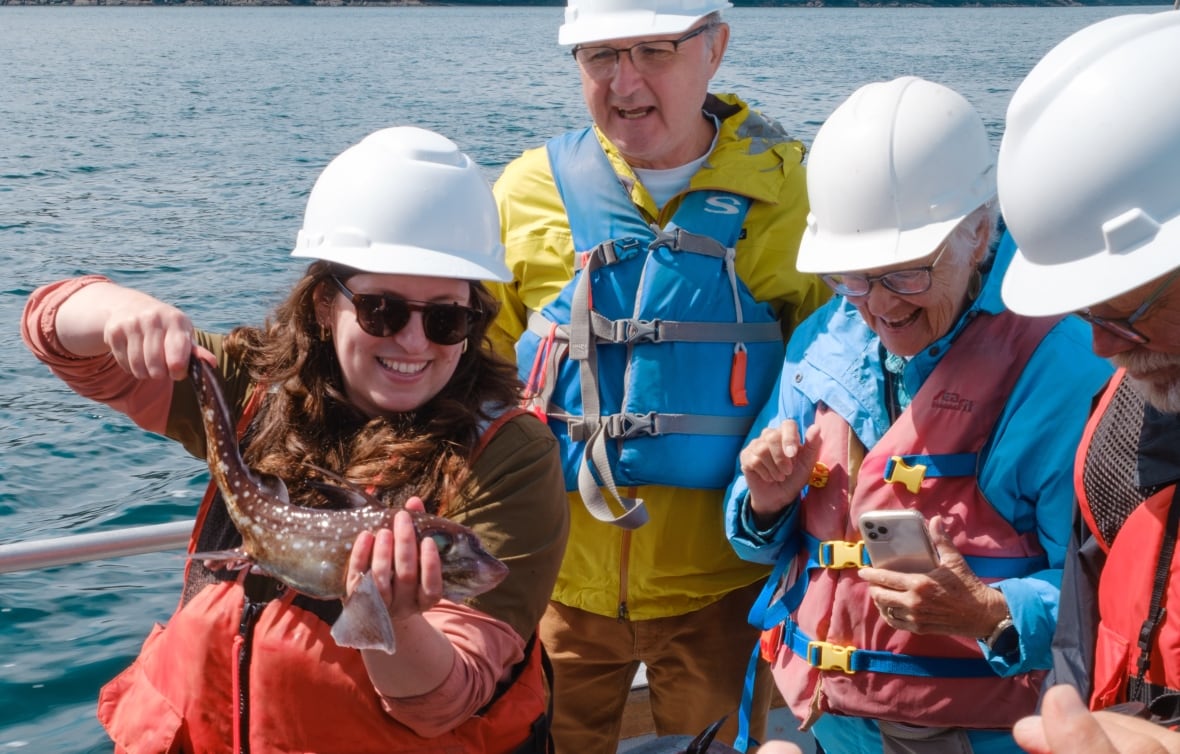 A smiling woman in a hard hat and sunglasses stands on a boat holding a long brown fish as others watch.