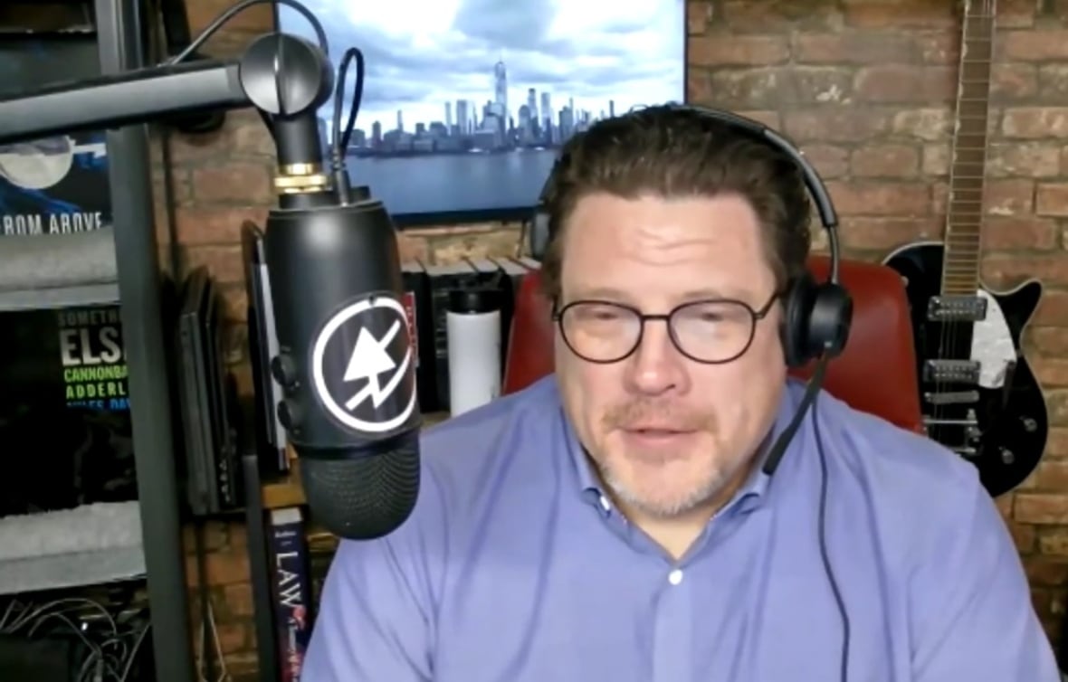 In a blue button up shirt and headphone a man sits in front of a microphone.