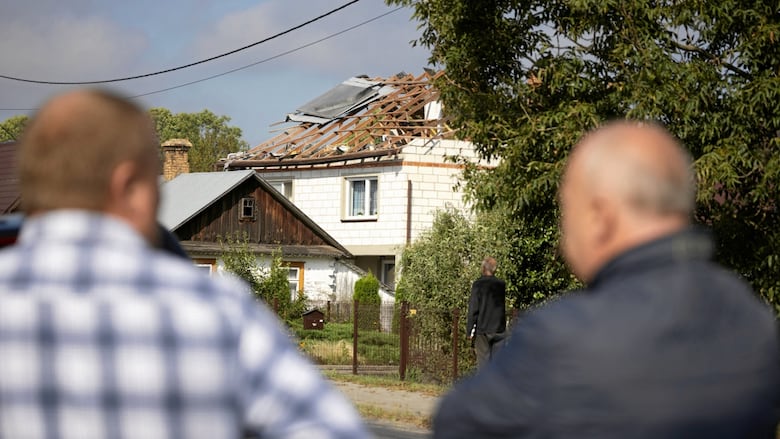 The backs of two men are in the foreground as they look toward a residential structure that has had its roof damaged, exposing wood columns.