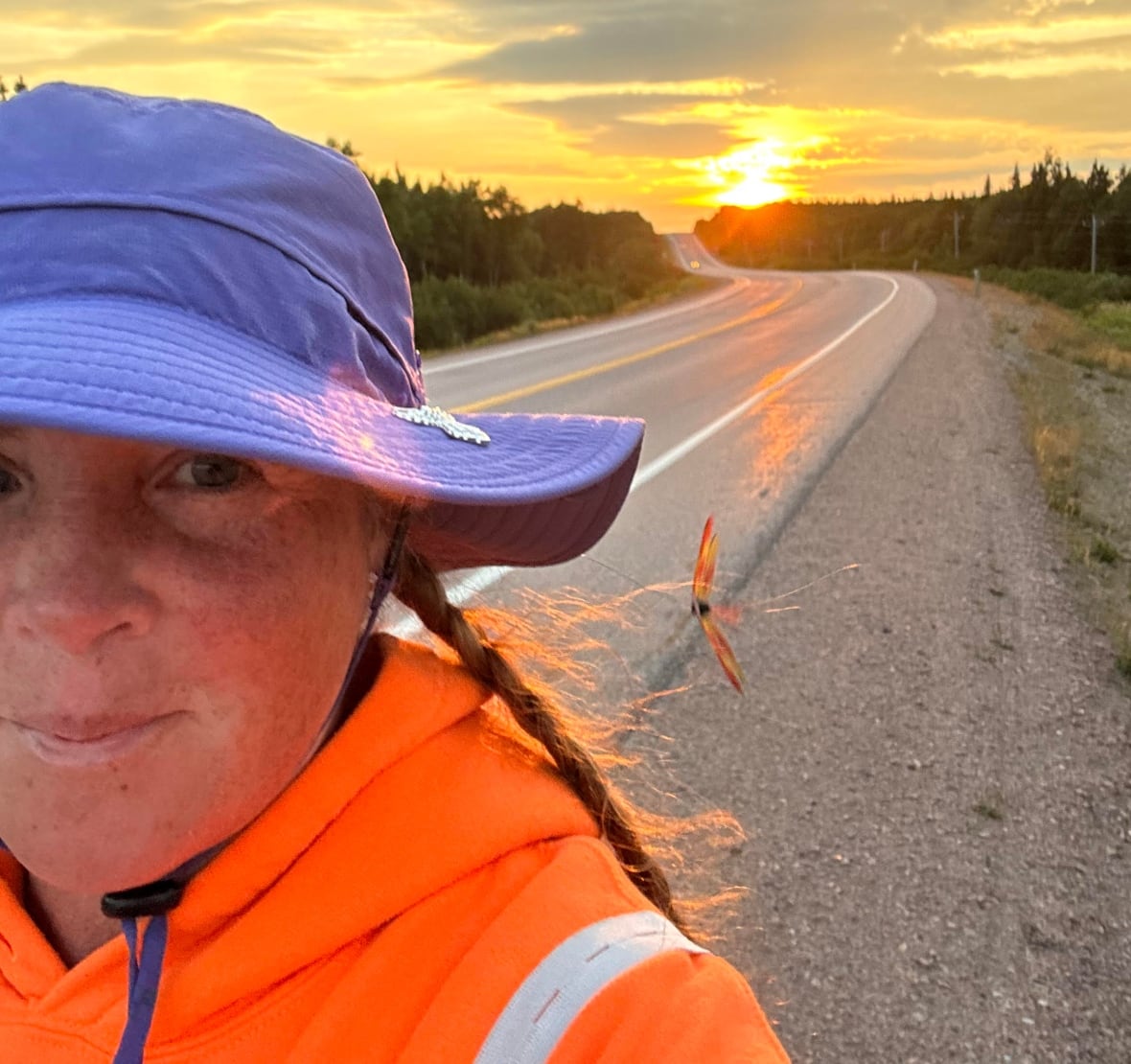 A woman wearing a purple hat and orange jacket walks on the road with the sun rising on the horizon.