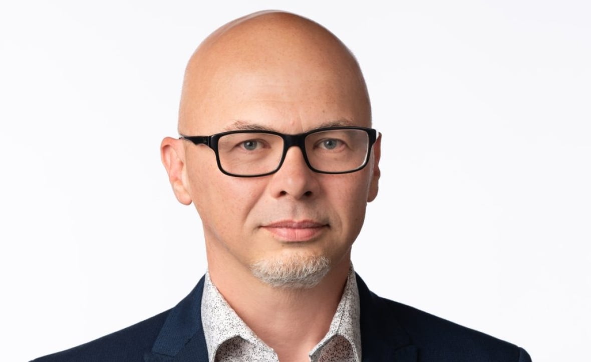 A man with a shaved head, black glasses and a suit jacket looks at the camera with a serious face. 