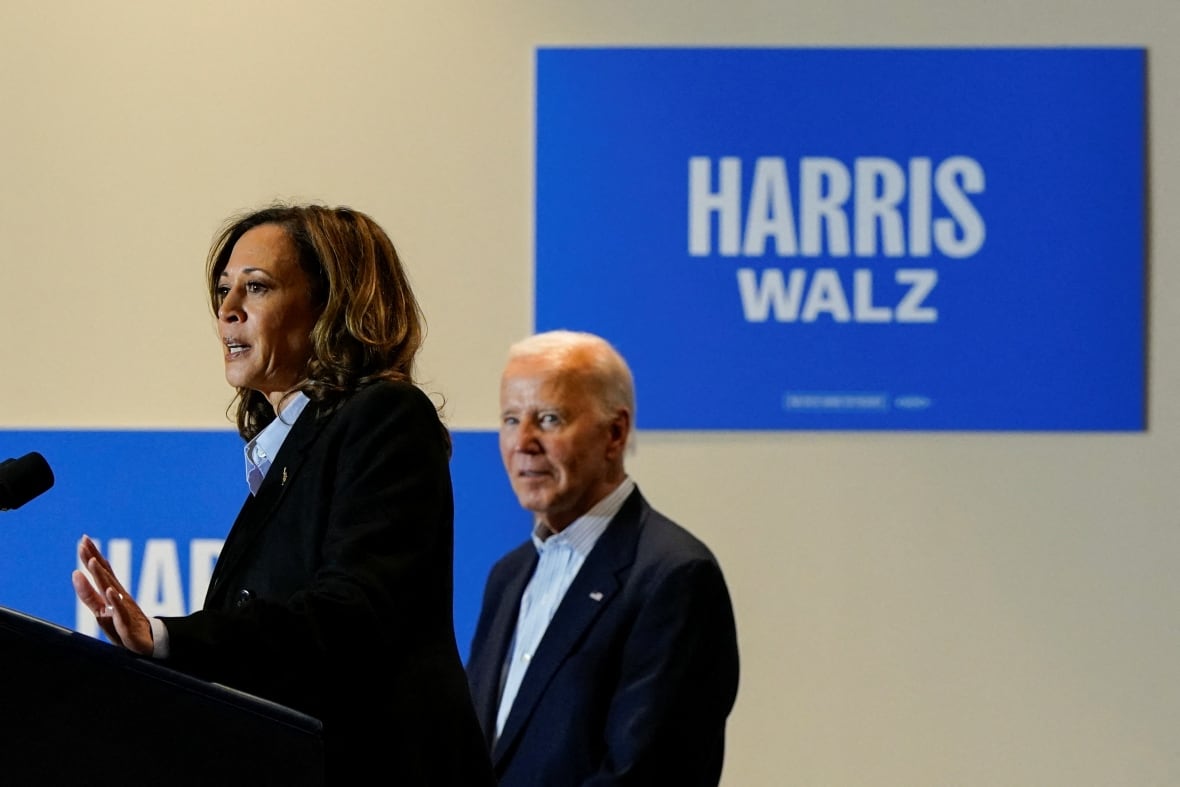 A woman with dark shoulder length hair speaks at a podium as a white haired clean shaven man in a suit jacket and collared shirt looks on.