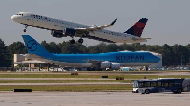 A blue plane that says "KOREAN" on the side sits on a runway while another plane takes off near it. 